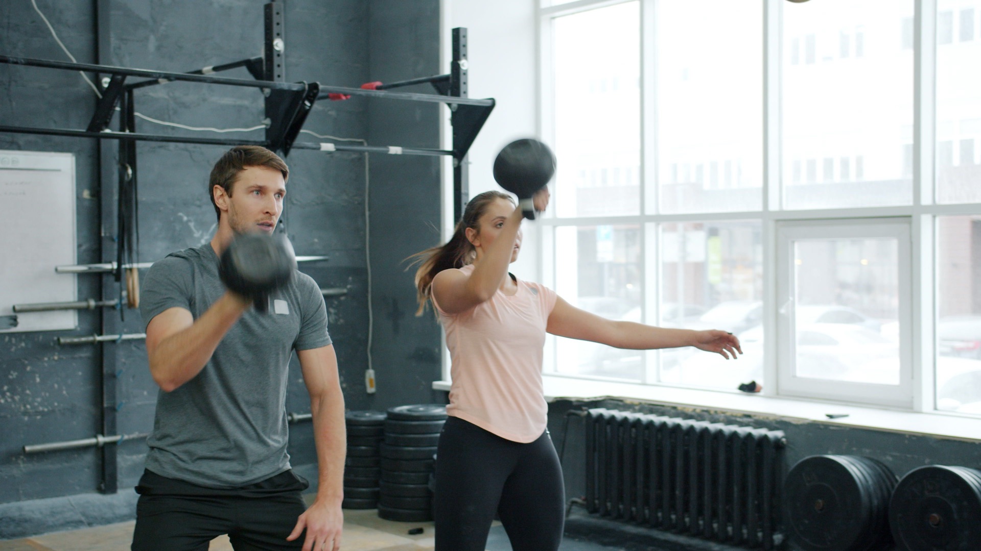 A man and a woman swinging kettlebells side by side in a bright gym, both looking straight ahead and concentrating.