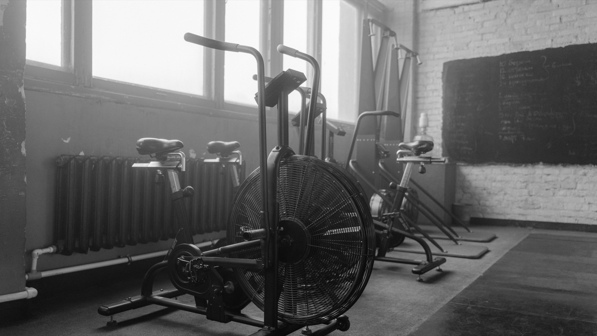 An empty gym in soft window light, a row of air bikes waiting, in black and white.