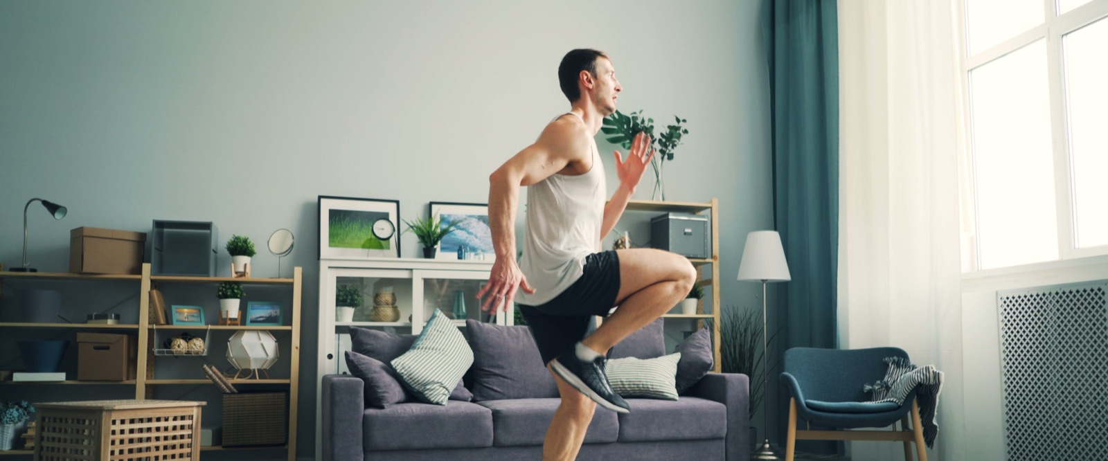 A man running on the spot in the middle of his living room, the sofa and a bright window behind him.